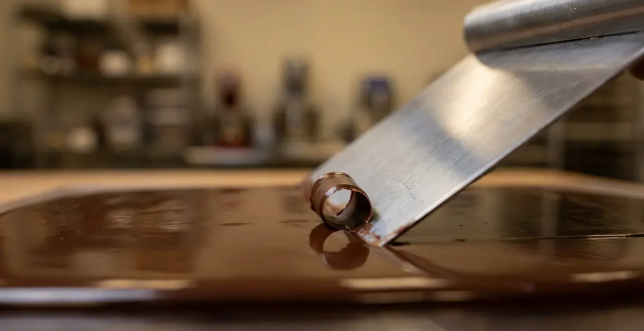 Close-up of metal bench scraper removing excess chocolate from polycarbonate mold